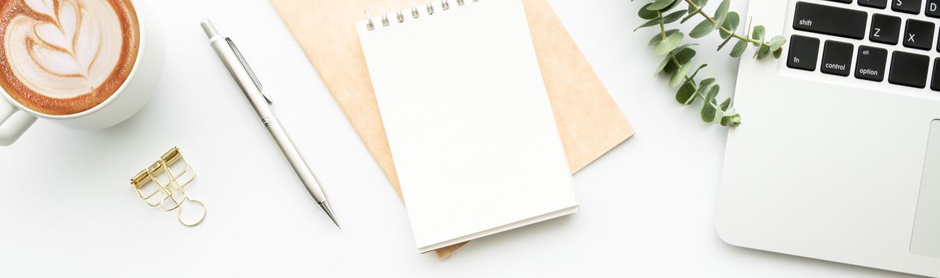 A flat lay of a workspace featuring a cup of coffee with latte art, a silver pen, a spiral notebook, a binder clip, some green leaves, and a portion of a laptop keyboard. Used as Image for Frequently Asked Questions