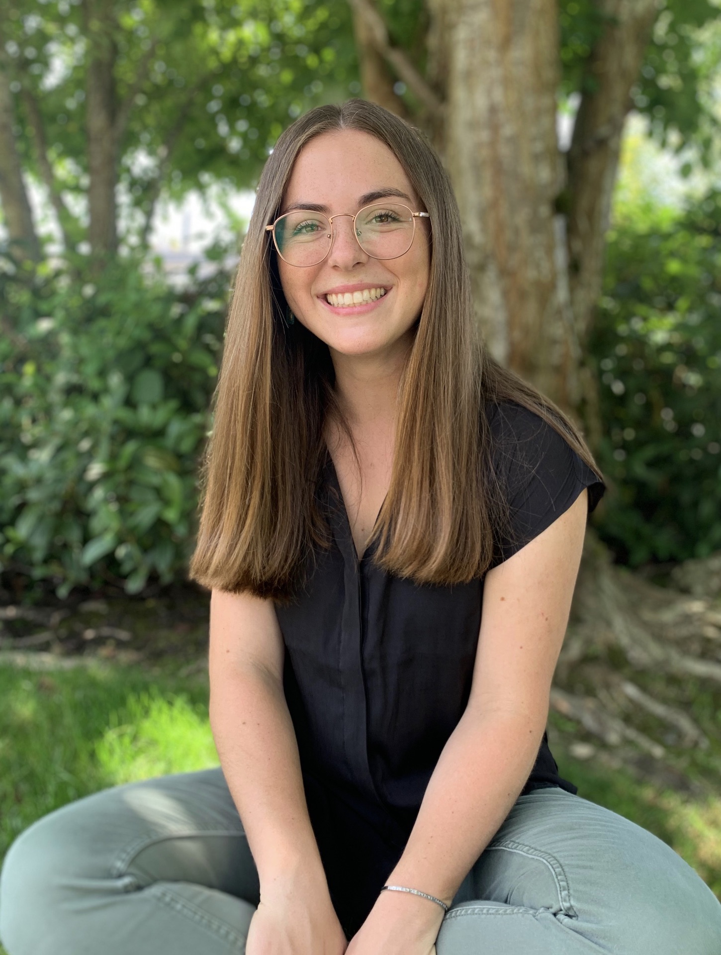 Young woman with glasses smiling, seated outdoors in front of a tree and greenery.